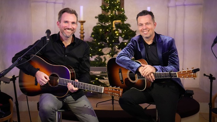 Mark og Christoffer sidder med guitarer og mikrofoner klar til julekoncert foran et pyntet juletræ.