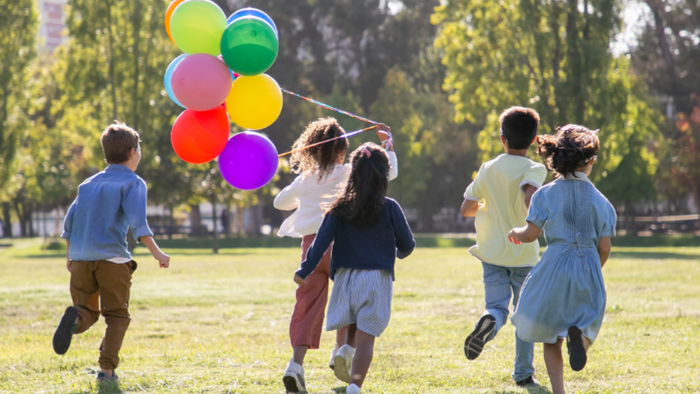 Kinder rennen fröhlich durch einen Park und jagen bunte Luftballons, die zusammengebunden sind.