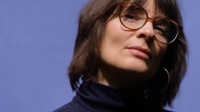 Eine Person mit kurzem Haar und Brille trägt einen dunklen Rollkragenpullover vor einem blauen Hintergrund.