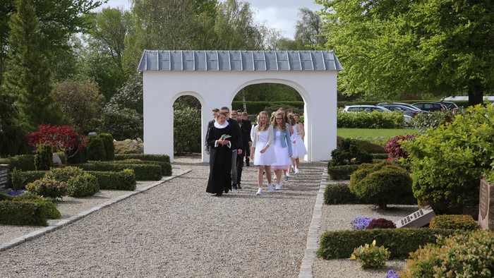 Et dejligt sommerbillede fra kirkegården, hvor præsten går forrest med konfirmander efter sig. De er på vej til gudstjenesten, hvor de skal konfirmeres.