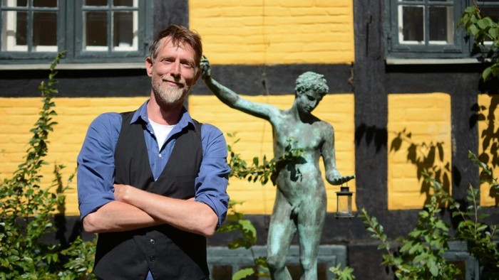 En mand poser selvsikkert ved siden af en klassisk statue foran en lysegul bygning med grønne planter.