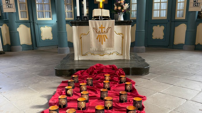 Elegante Altaraufstellung mit zahlreichen goldenen Votivkerzen auf einem roten Tuch, eingerahmt von einem dekorativen Kircheninnenraum.