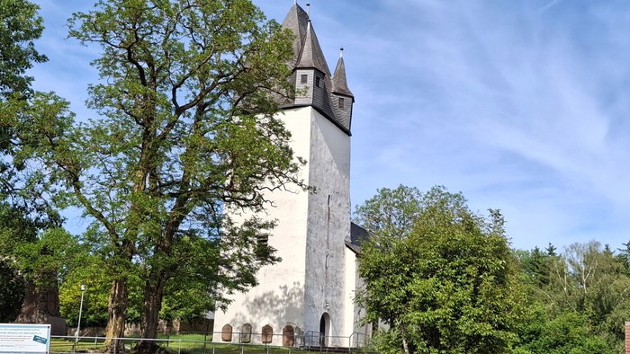 Historischer Steinkirchturm, umgeben von üppigem Grün unter einem klaren blauen Himmel.