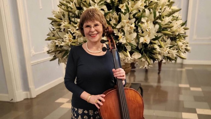 En kvinde i elegant påklædning holder en cello og står ved siden af en stor blomsterdekoration indendørs.