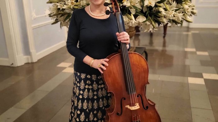 En kvinde i elegant påklædning holder en cello og står ved siden af en stor blomsterdekoration indendørs.