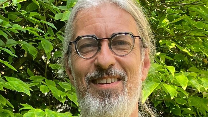 Ein lächelnder Mann mit Brille, langen Haaren und Bart, der zwischen grünem Laub steht.