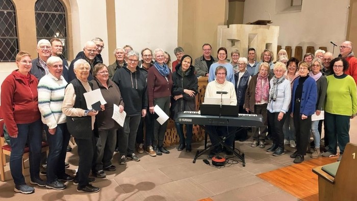 Gruppe von Menschen, die sich drinnen versammelt haben, wahrscheinlich ein Chor oder eine Gemeinschaftsveranstaltung, posieren mit Notenblättern und einem Pianisten.