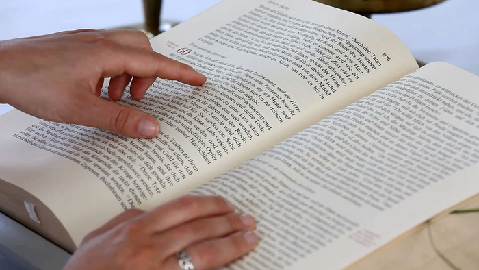 Eine Hand zeigt auf eine aufgeschlagene Buchseite, während die andere Hand auf dem Buch liegt.