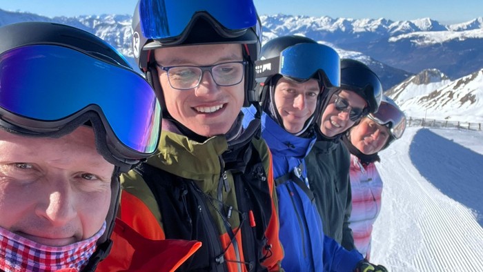 Gruppe von Skifahrern auf einem sonnigen Berghang, die Helme, Skibrillen und Winterkleidung tragen und fröhlich zusammen posieren.
