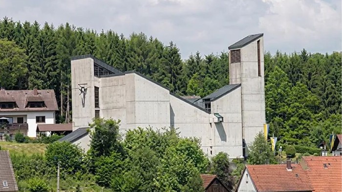 Moderne Betonkirche, umgeben von üppigem Grün und Wohnhäusern an einem Hang.