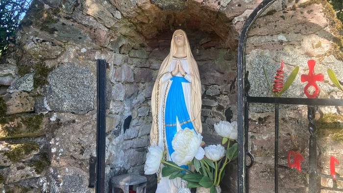 Statue der Jungfrau Maria in einer steinernen Nische mit Blumenspenden und religiösen Symbolen in der Nähe.