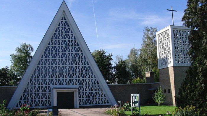 Moderne dreieckige Kirche mit geometrischem Gitterdesign und angrenzendem Glockenturm, umgeben von gestalteter Grünanlage.
