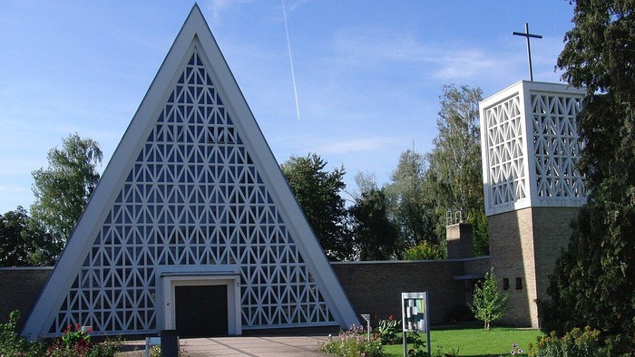 Moderne dreieckige Kirche mit geometrischem Gitterdesign und angrenzendem Glockenturm, umgeben von gestalteter Grünanlage.