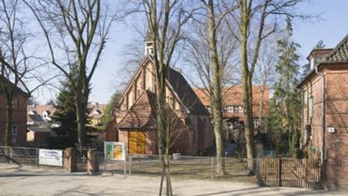 Ein Kirchengebäude mit hohem Dach und umgebenden Bäumen, daneben ein Parkplatz mit wenigen Autos.