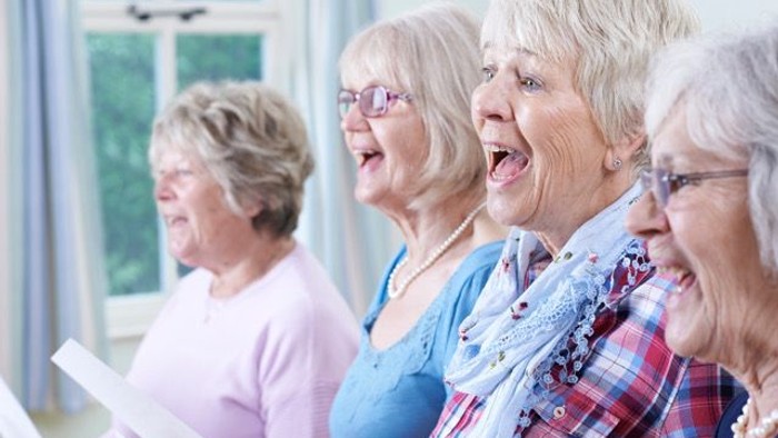 Ein Gruppe älterer Frauen singt gemeinsam in einem Raum mit großen Fenstern.