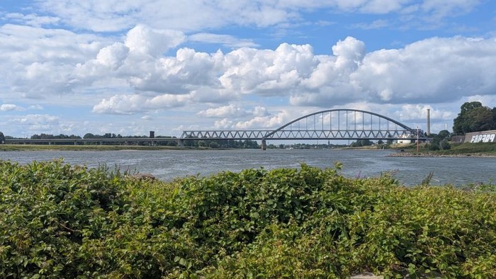 Ein malerischer Blick auf einen breiten Fluss mit einer großen Bogenbrücke unter einem teilweise bewölkten Himmel.