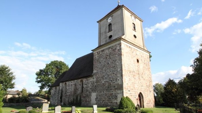 Historische Steinkirche mit hohem Glockenturm in einem ruhigen, grasbewachsenen Friedhof.