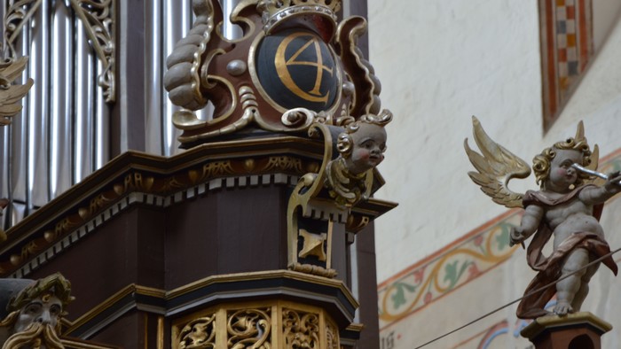Komplekst orgel med udsmykkede udskæringer og en englefigur, der holder et kors i en kirkes indre.