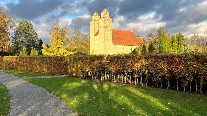 Hyggelig kirke gemt i frodig grønne omgivelser ved siden af en brostensbelagt sti under en dramatisk himmel.