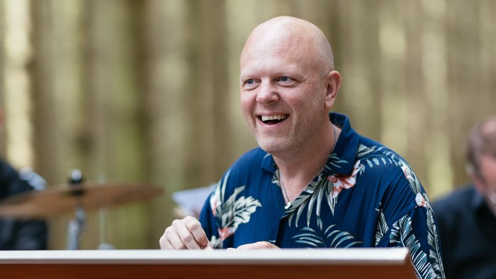 Et band optræder udendørs med en skaldet mand, der spiller på et keyboard og smiler, mens andre spiller trommer og guitar.