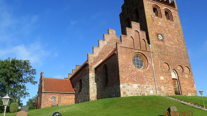 En middelalderkirke med tårn og gravsteder på en grøn bakke