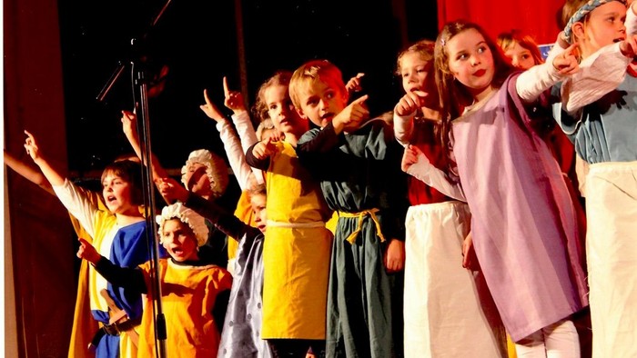 Kinder treten in bunten Kostümen auf der Bühne auf und heben begeistert die Arme während eines Schultheaterstücks.