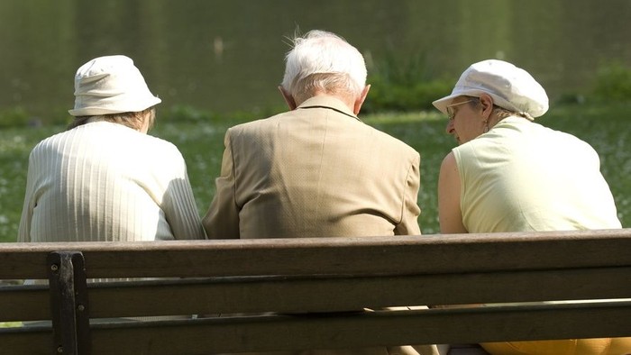 Drei ältere Menschen sitzen auf einer Parkbank und schauen in die Ferne.
