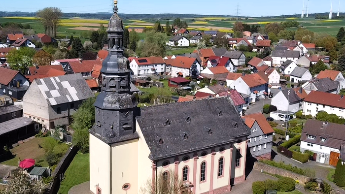 Luftaufnahme eines malerischen Dorfes mit einer markanten Kirche, umgeben von Häusern mit roten Dächern und grünen Feldern.