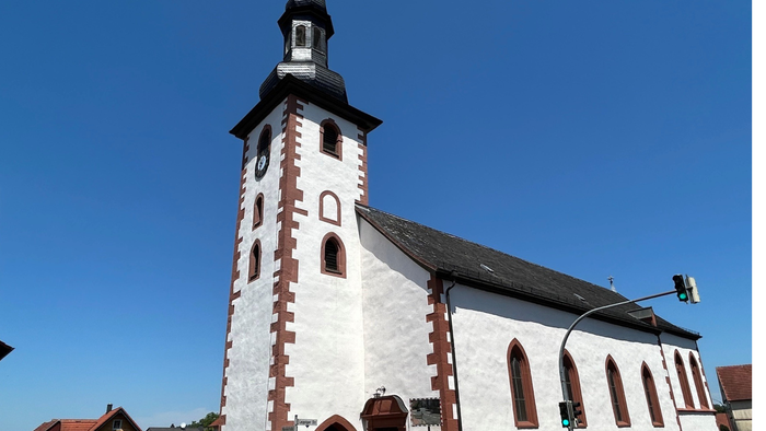 Eine charmante weiße Kirche mit einem hohen Glockenturm und roten Backsteinakzenten steht unter einem klaren blauen Himmel.