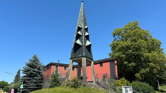 Eine markante Kirche mit einem hohen, pyramidengekrönten Turm, die sich vor einem strahlend blauen Himmel und üppigem Grün abhebt.