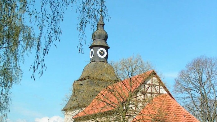 Kleine Kirche mit rotem Dach und Uhrturm, umgeben von grünem Rasen und Bäumen.