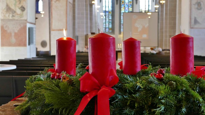 Das Foto zeigt den Adventskranz in der Stiftskirche Kaufungen mit einer angezündeten Kerze. Im Hintergrund die Pfeiler und Fenster der Kirche und die Leinwand für Fotos von der Aktion
