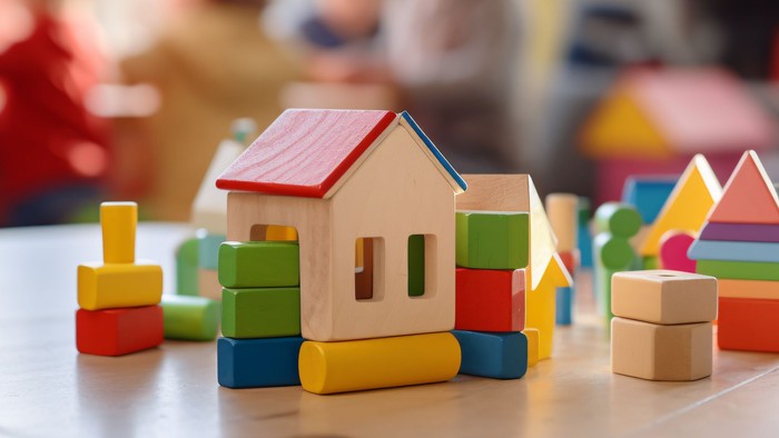 Bunte Holzklötze, zu einem kleinen Haus auf einem Kindertisch im Klassenzimmer arrangiert, mit Kindern im Hintergrund.