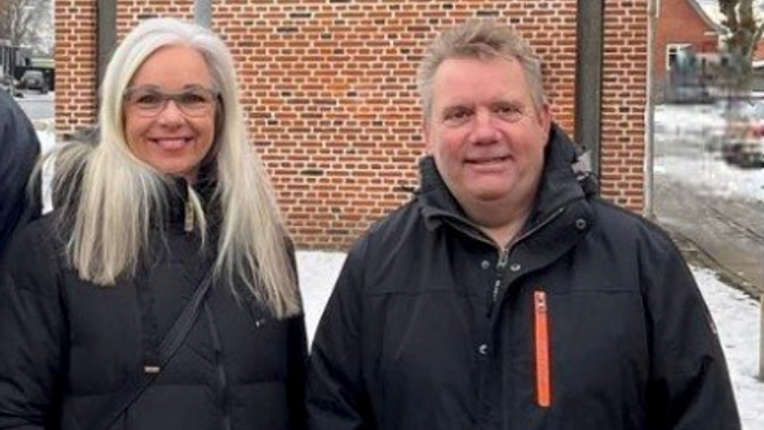 To smilende personer i vinterjakker, der står udendørs nær en murstensbygning.