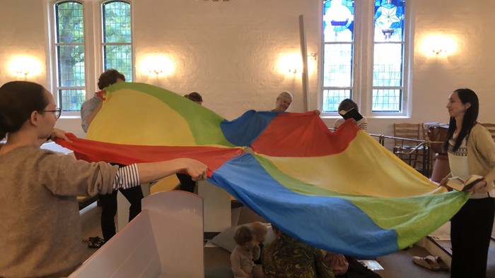 Mennesker holder glædeligt og snurrer et stort regnbuefarvet faldskærm indendørs under en gruppeaktivitet.