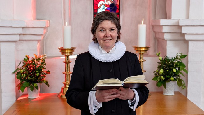 En person i religiøst tøj holder en åben bog og smiler i en kirkelig omgivelse med lys og blomster.