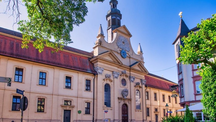 Historische Kirche mit Uhrturm und aufwendiger Fassade, umgeben von Bäumen und anderen Gebäuden.