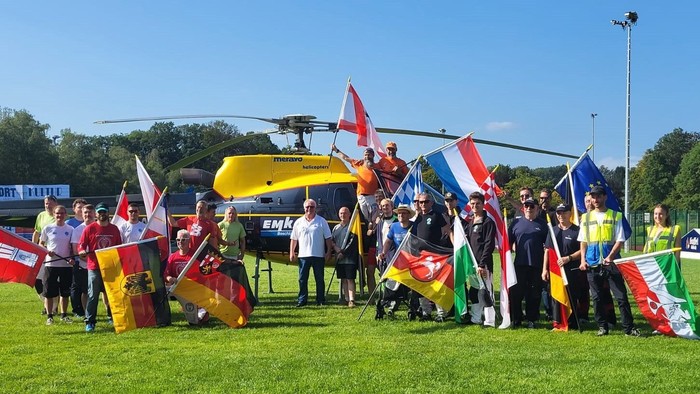 Gruppe mit bunten Flaggen neben einem gelben Hubschrauber auf einem grasbewachsenen Feld während einer Veranstaltung.
