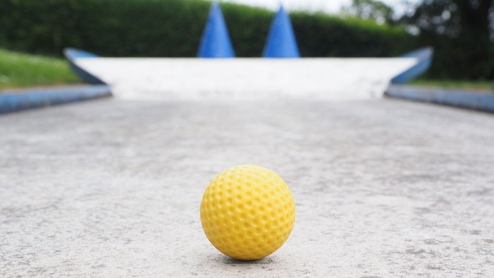 Eine leuchtend gelbe Golfball liegt auf einer glatten, strukturierten Putt-Grün-Oberfläche mit blauen Ausrichthilfen im Hintergrund.