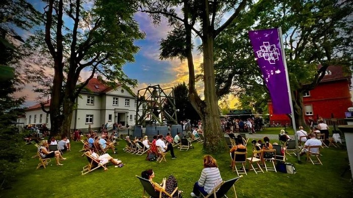 Menschen sitzen an einem Sommerabend auf einer Wiese in Liegestühlen unter großen Bäumen. Im Hintergrund stehen Gebäude und eine Bühne, daneben eine violette Fahne mit Facettenkreuz. Die Atmosphäre wirkt entspannt und gemeinschaftlich.