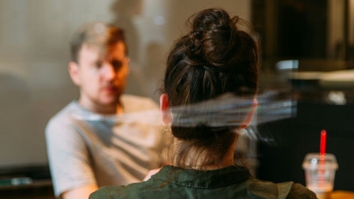 Zwei Personen sitzen in einem schwach beleuchteten Raum, eine von ihnen abgewandt mit hochgestecktem Haar zu einem Dutt.