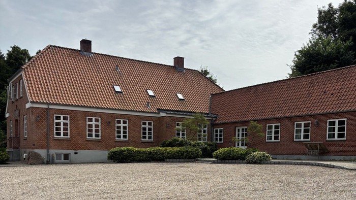 Et murstensbygning med rødt teglstenstag omgivet af en grusgård og anlagt beplantning.