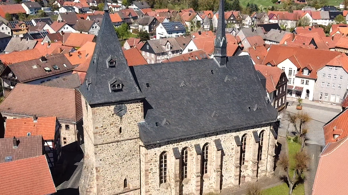 Luftaufnahme einer historischen Steinkirche, umgeben von malerischen Dorfhäusern mit roten Dächern.