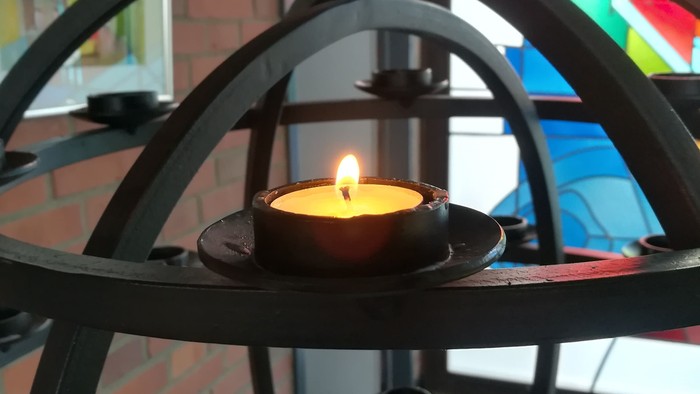 Eine brennende Kerze in einem schwarzen Metallhalter vor einem bunten Glasfenster