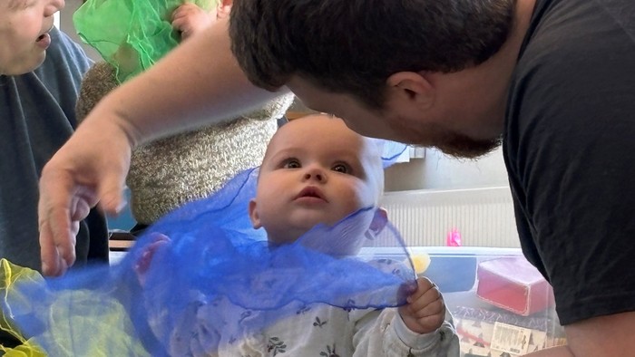En mand hjælper en baby, der sidder på et farverigt tæppe med en blå klud i nærheden.