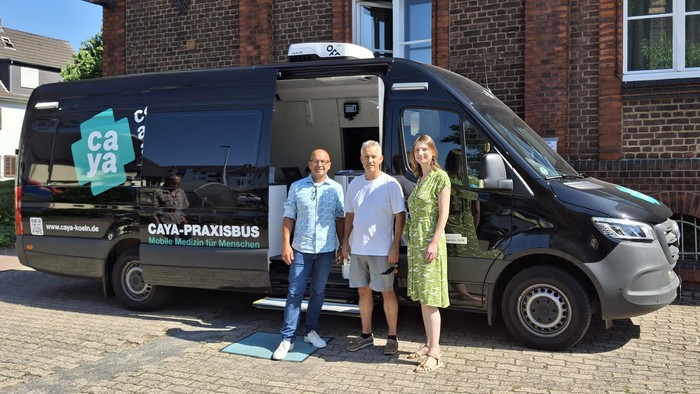 Drei Personen stehen neben einem schwarzen „Caya-Praxisbus“-Medizintransporter vor einem Backsteingebäude.