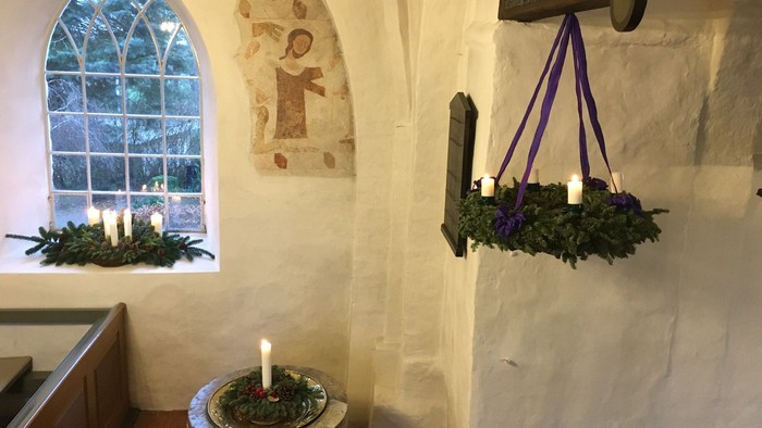 Kirkerum med adventskrans hængt op med lilla bånd og to andre juledekorationer i hhv. vindueskarm og på døbefont.