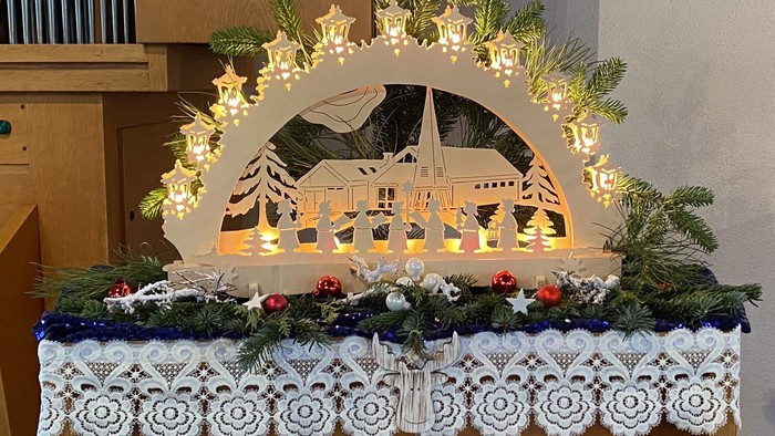 Elegante Weihnachtskranz-Dekoration mit beleuchteter Weihnachtskrippe, Tannenzweigen und festlichen Schmuckelementen auf einer gehäkelten Tischdecke.