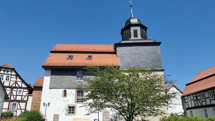 Das Bild zeigt eine Kirche mit einem Turm und rotem Dach, umgeben von grünen Büschen und Bäumen