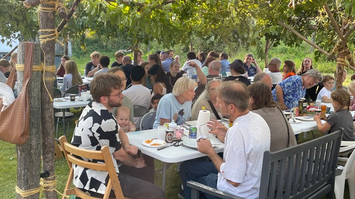 En gruppe af mennesker sidder ude og spiser på bord og benker under træer.